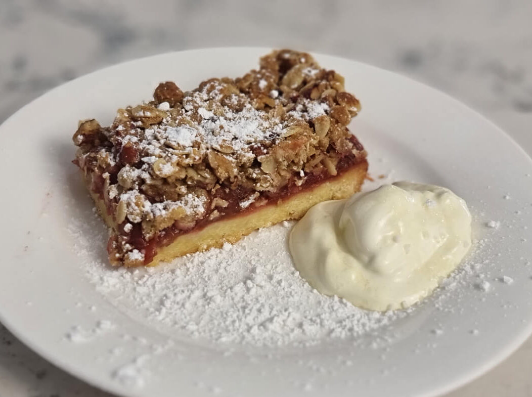 A piece of rhubarb crumble slice with a dollop of sour cream, sprinkled with icing suagr